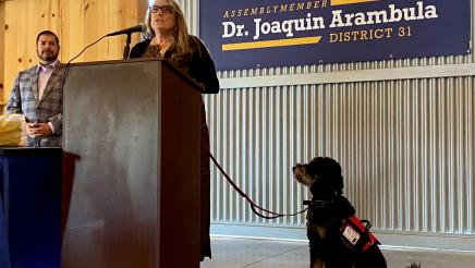 Dr. Kathleen “Kathi” Rindahl speaks to the audience at the March 25, 2022 luncheon. She is Assemblymember Arambula’s honoree for the 2021 award. With her is her service dog, Riley.